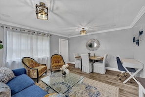 A living room with a blue sofa, two patterned armchairs, a glass coffee table, and a dining area with a wooden table and white chairs.