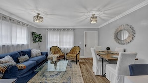 Ein Wohnzimmer mit einem blauen Sofa, zwei Sesseln, einem Glastisch, einem Esstisch mit weißen Stühlen und einem dekorativen Spiegel an der Wand.