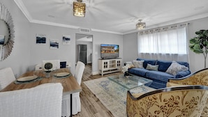 Ein modernes Wohnzimmer mit einem Glastisch, einem blauen Sofa und einem Essbereich mit einem Holztisch, gedeckt für zwei Personen.