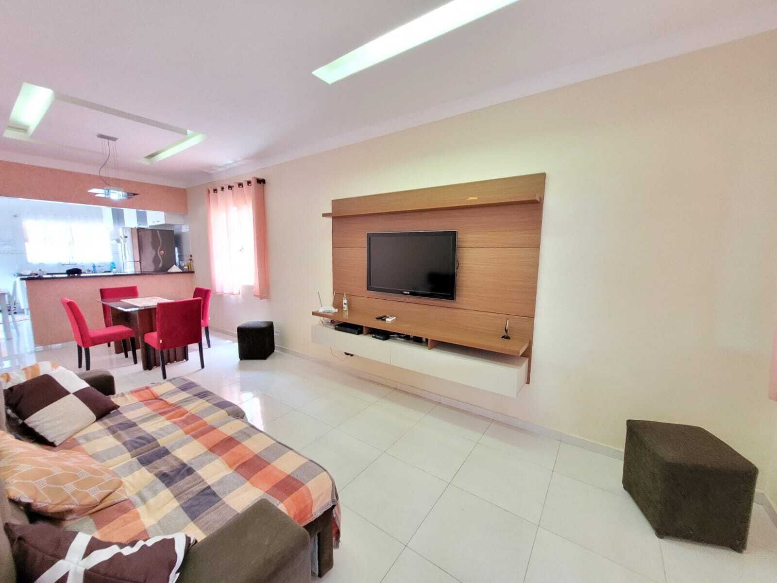 A modern living room with a sofa, a TV mounted on the wall, a dining area with chairs, and a kitchen in the background.