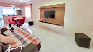 Una sala moderna con un sofá, un televisor en la pared, un comedor con sillas y una cocina al fondo.