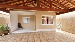 Un porche cubierto con suelo de piedra, vigas de madera y vistas a una habitación interior con una silla y un ventanal.