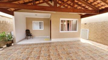 A covered patio with stone flooring, wooden beams, and a view of an interior room with a chair and a window.