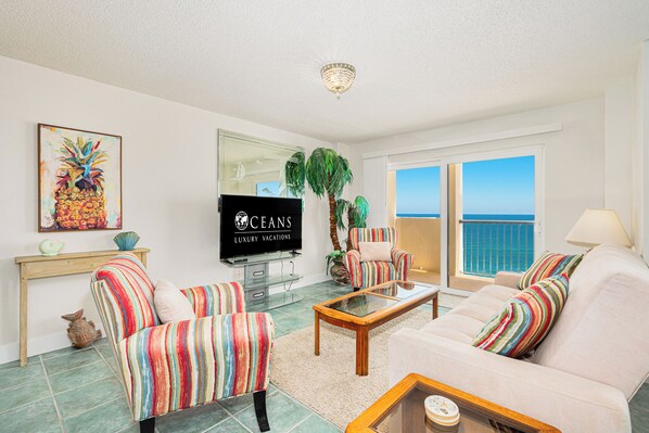 A living room with striped armchairs, a sofa, a coffee table, a TV, and a large window with a view of the ocean.