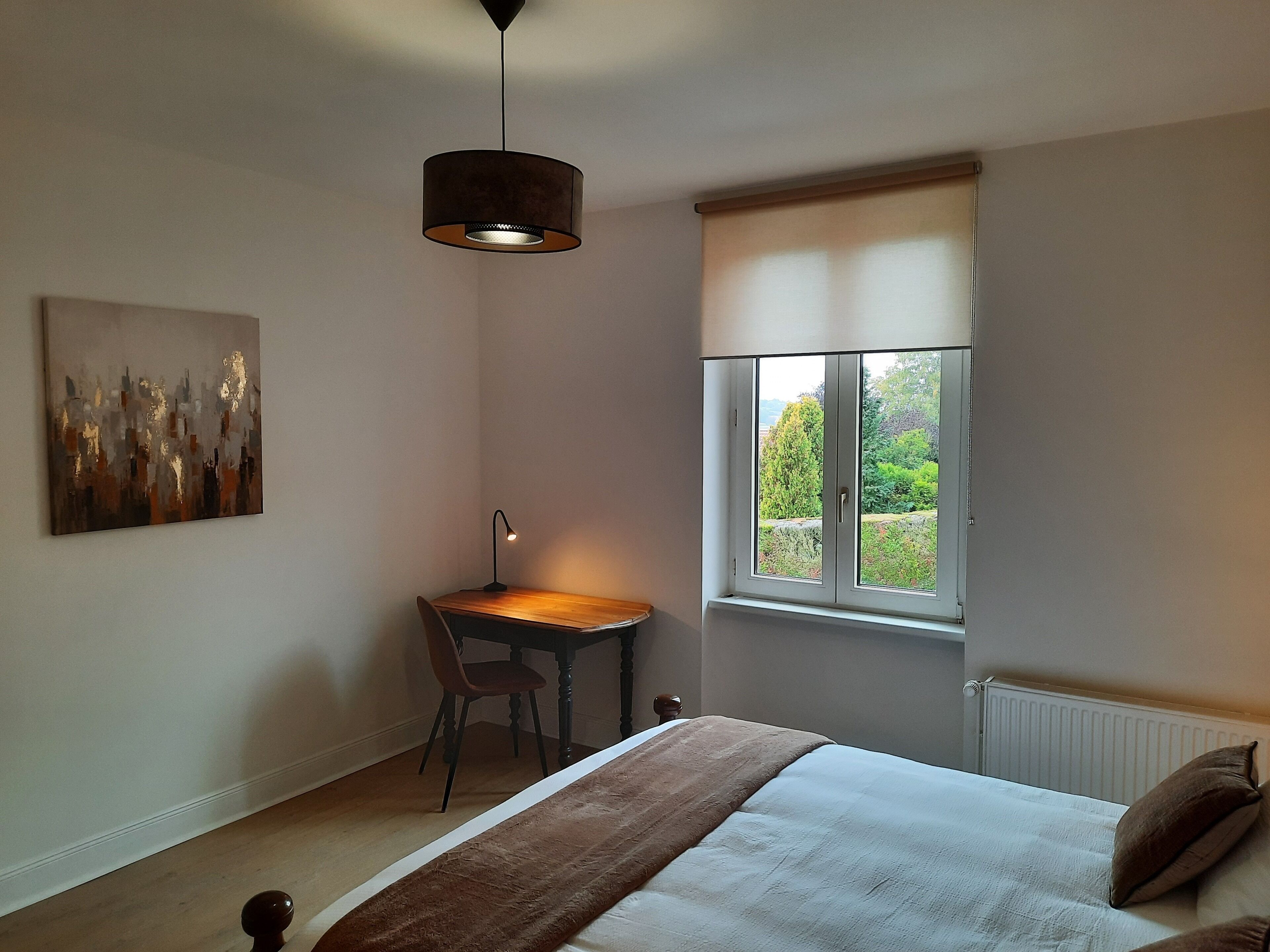 A bedroom with a bed, a desk, a chair, a lamp, a window with a view of greenery, and a painting on the wall.