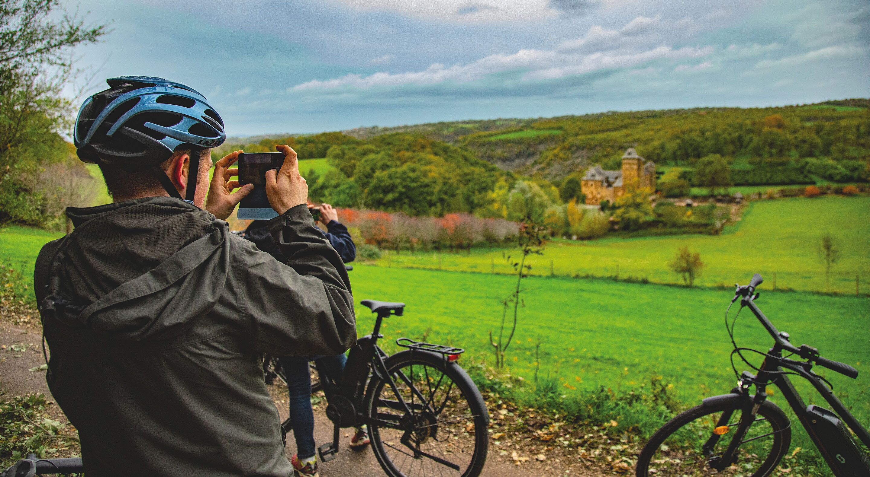 A person taking a photo with a camera, a bicycle, and a scenic view of a green landscape with a building in the distance.