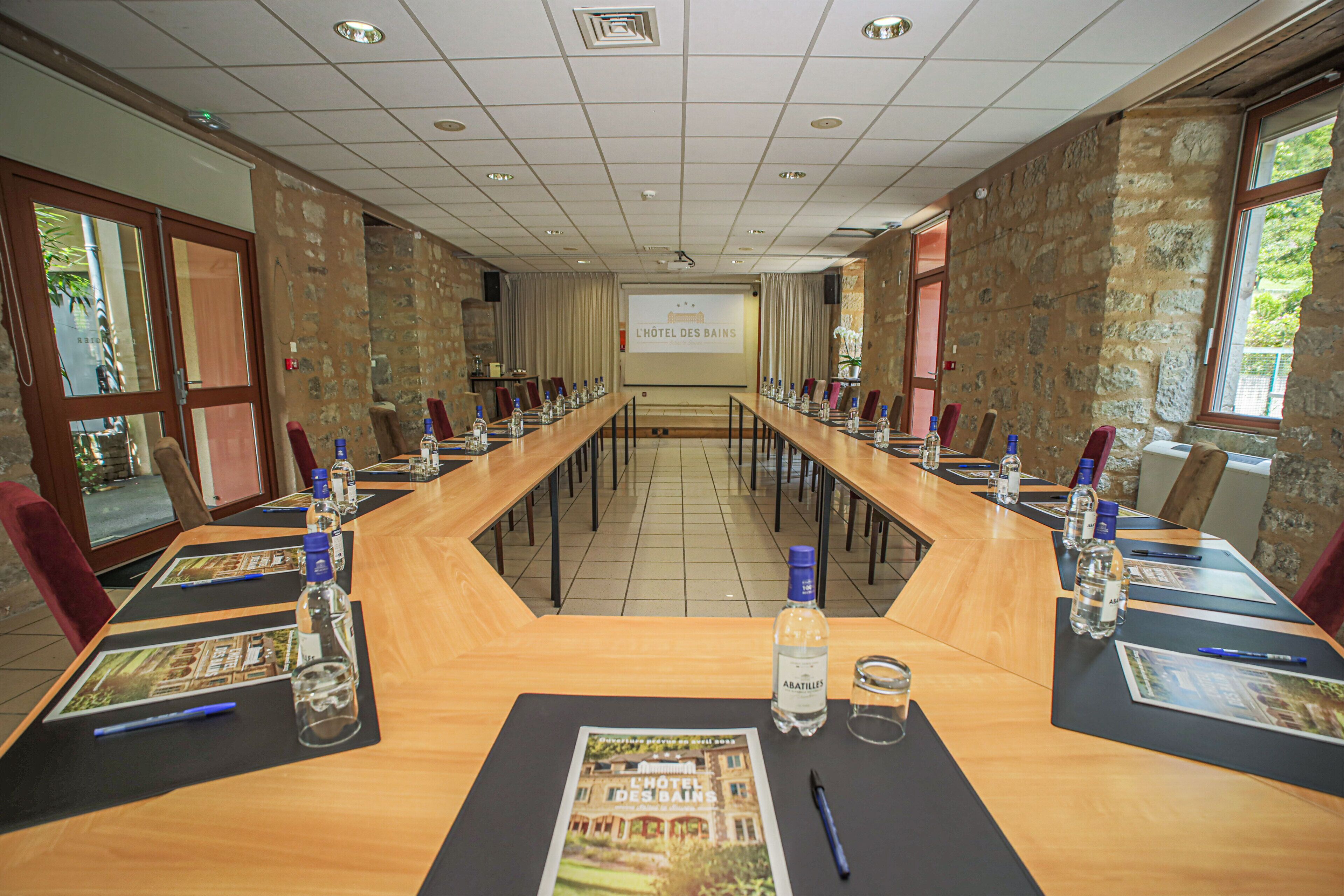 A conference room with a long table, chairs, water bottles, and a projector screen displaying "L'Hôtel des Bains".