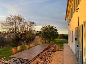 An outdoor dining area with a long wooden table and chairs, surrounded by trees and a building with shutters.