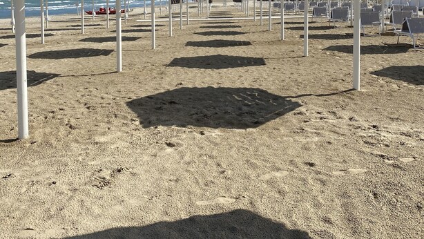 Ein Strand mit weißen Sonnenschirmen und Liegestühlen, ein Boot im Wasser und ein klarer Himmel.