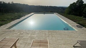 Ein rechteckiger Swimmingpool mit glasklar blauem Wasser, umgeben von einer Steindeck. Zwei Holzliegen sind zum Pool hin ausgerichtet.