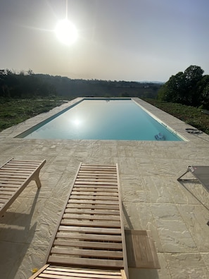 A rectangular swimming pool with a clear blue water surface, surrounded by a stone deck. There are two wooden lounge chairs facing the pool.