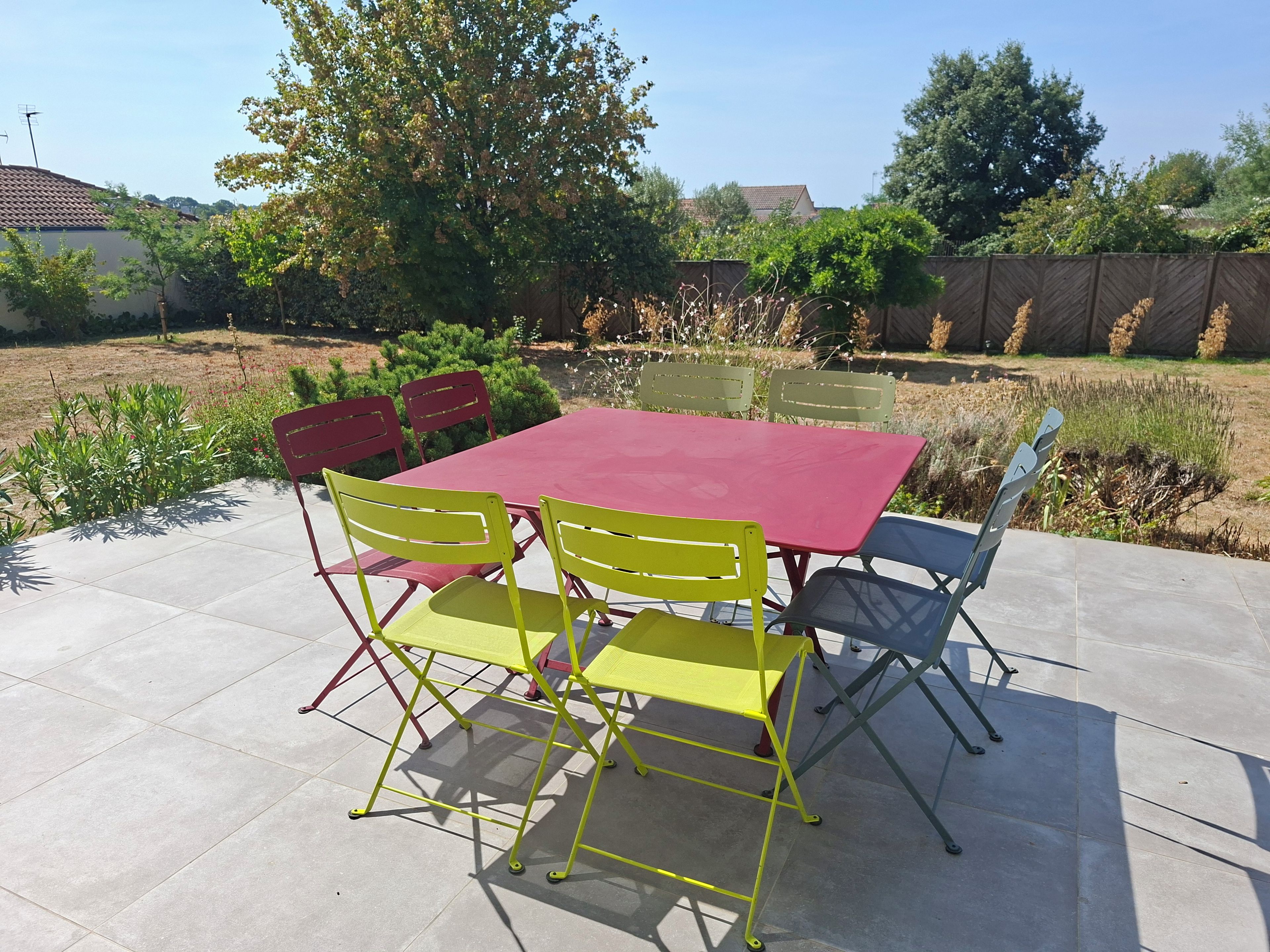 Een buitenpatio met een rode tafel en stoelen, omgeven door groen en een hek.