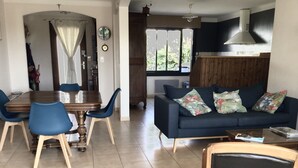 A dining area with a wooden table and blue chairs, a sofa with floral cushions, and a kitchen area with a stove and window.