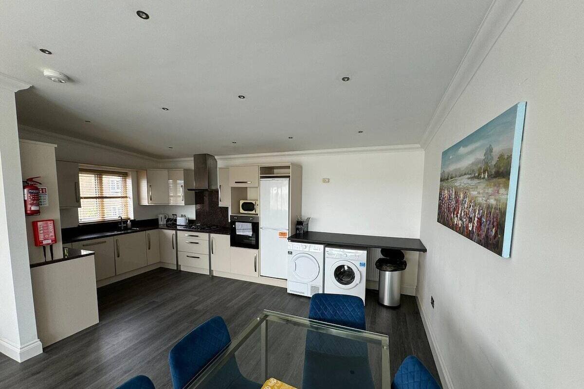 A modern kitchen with white cabinets, a microwave, and a washing machine. There is a glass dining table with blue chairs.