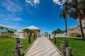 A pathway leading to a building with a flat roof, surrounded by palm trees and a clear blue sky.