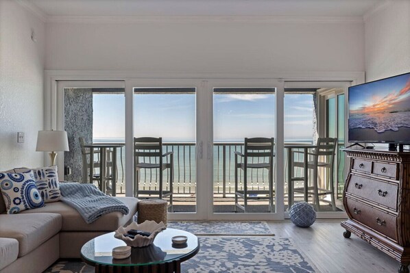 A living room with a sofa, coffee table, and TV, leading to a balcony with chairs and a view of the ocean.