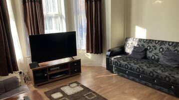 A living room with a flat-screen TV on a wooden stand, a sofa with floral upholstery, and a brown rug with geometric patterns.