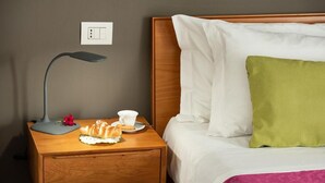 Ein ordentlich bezogenes Bett mit weißen und grünen Kissen, ein Nachttisch aus Holz mit Lampe, Tasse und Teller mit Speisen.