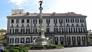 Ein großes Gebäude mit einem zentralen Denkmal, das eine Figur auf einer Säule zeigt, umgeben von kleineren Statuen und einem Pflasterplatz.