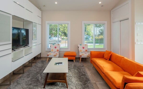 A modern living room with a white sofa, an orange sofa, a coffee table, and two chairs with colorful patterns. - Modern and cool cottage close to pebble beach! (Middletown)