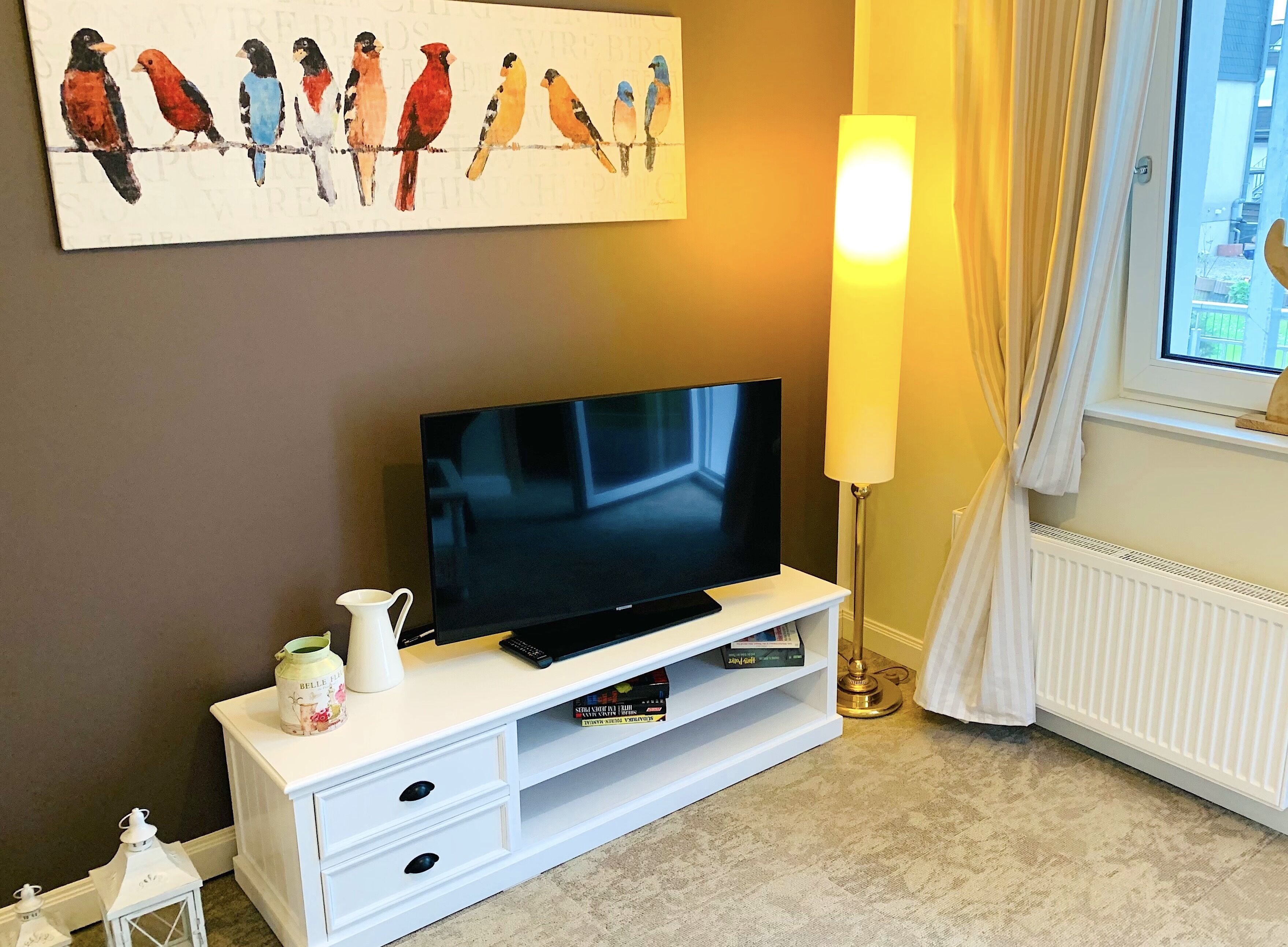 Sala de estar com televisão de tela plana em um móvel branco, luminária de chão, pintura de pássaros na parede e janela com cortinas.