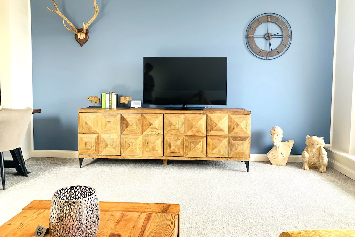 A living room with a blue wall, a mounted deer head, a clock, a wooden cabinet with decorative patterns, and a television.