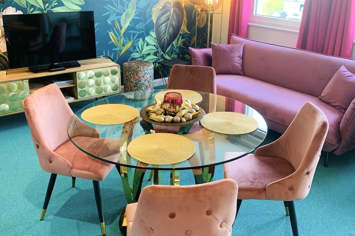 A living room with a glass-top dining table surrounded by pink upholstered chairs, a sofa, and a television on a stand.