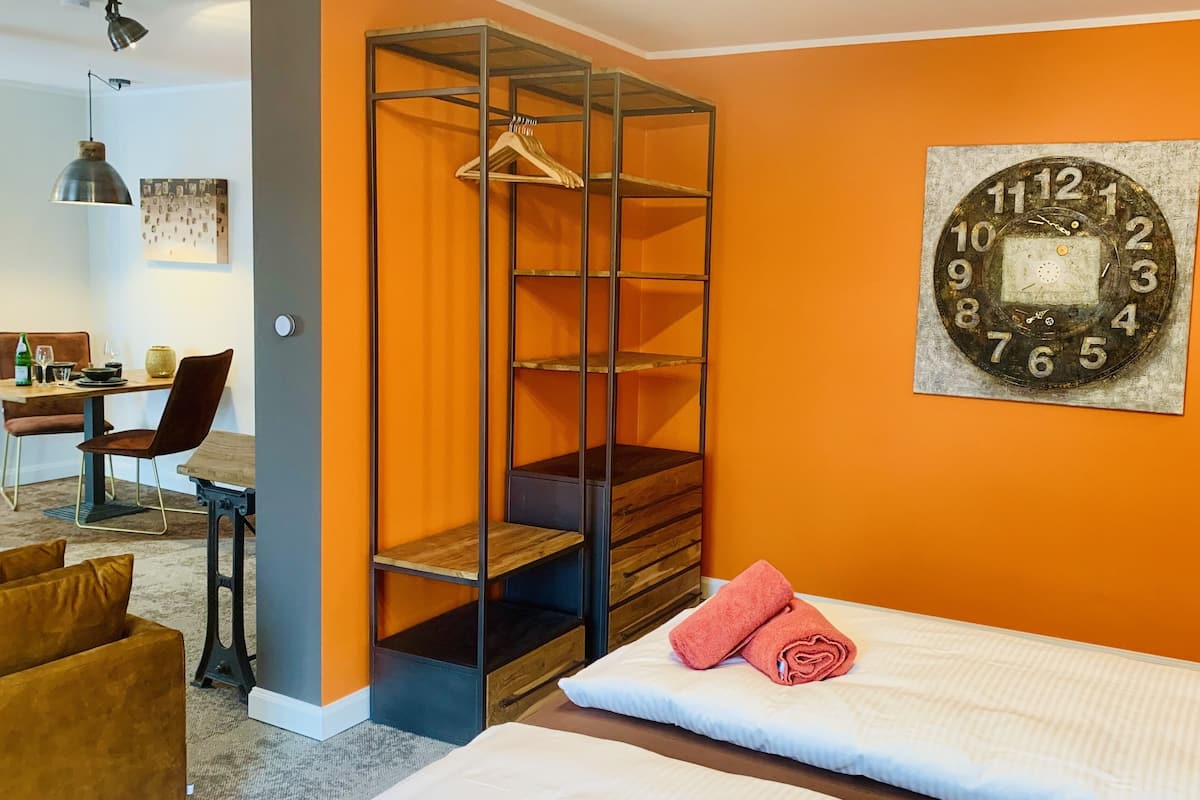 A bedroom with a bed, a hanging wardrobe, a clock on the wall, and a dining area in the background.