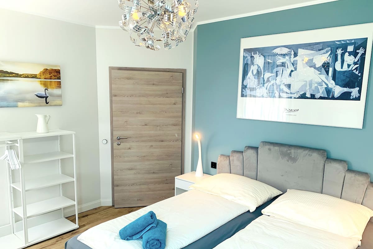 A modern bedroom with a wooden door, a bed with white bedding and blue towels, a white shelving unit, and a Picasso painting on the wall.