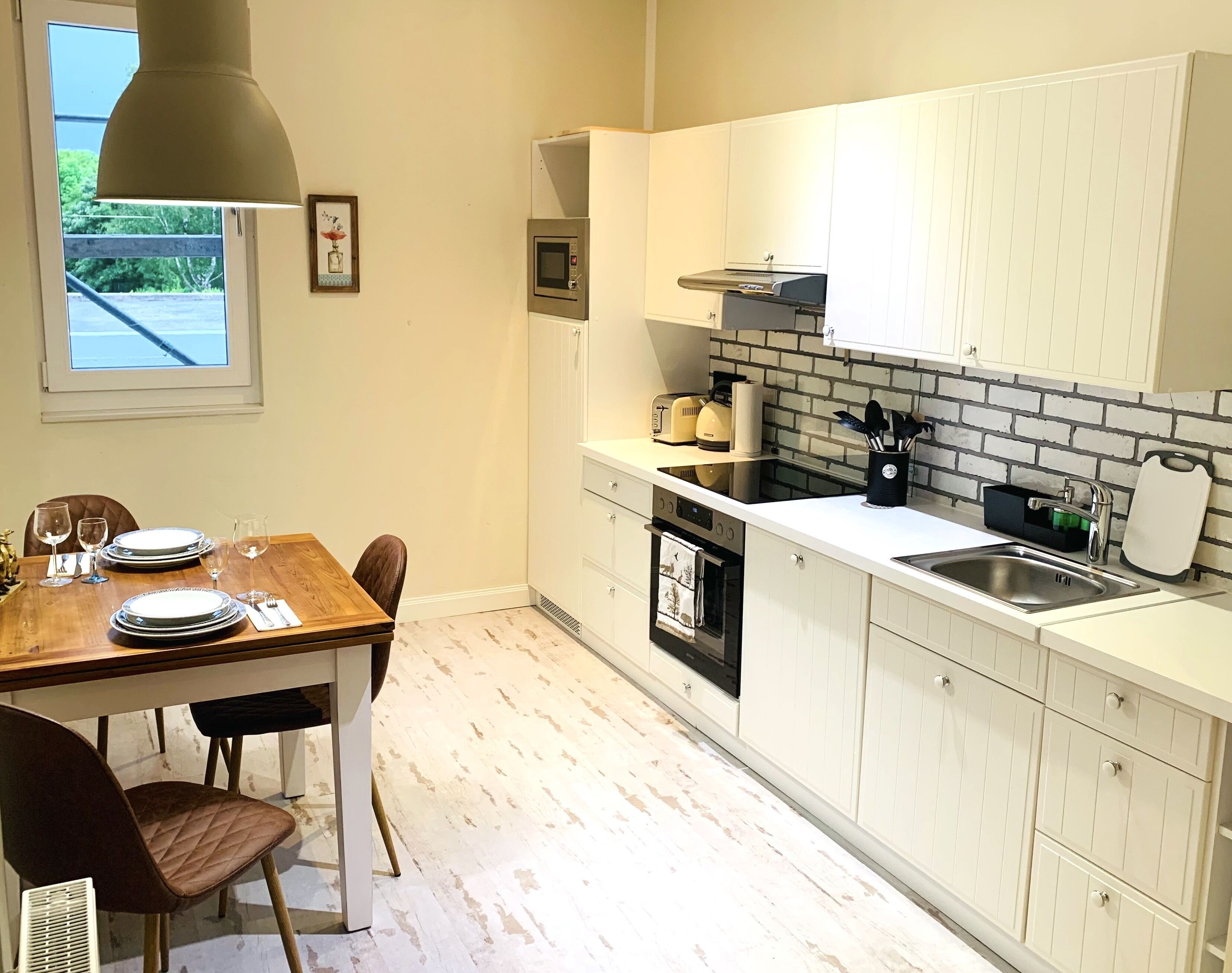 A kitchen with white cabinets, a brick backsplash, a dining table set with plates and glasses, and a window with a view of greenery.