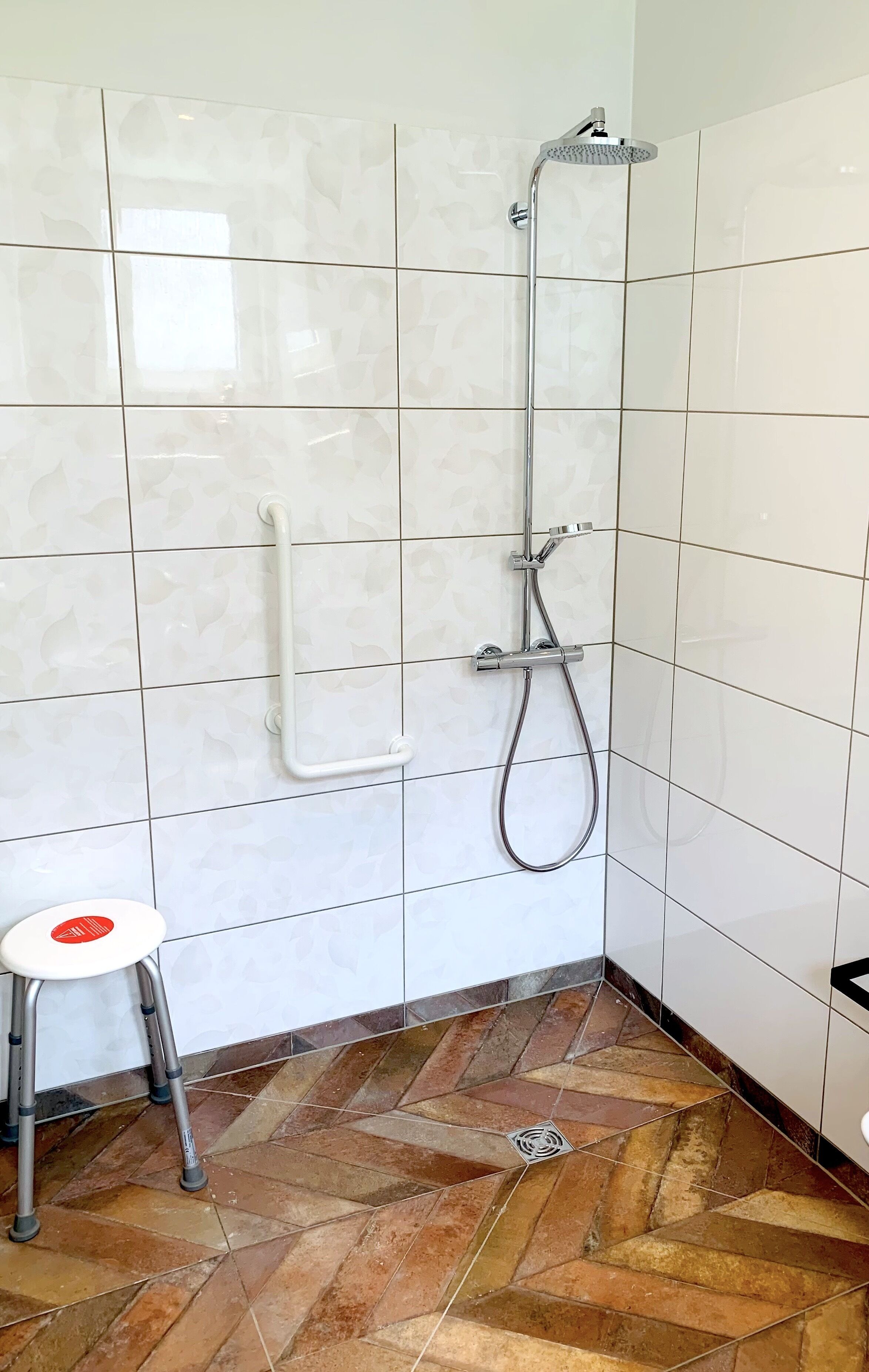 A bathroom with white tiled walls, a shower with a handheld showerhead, and a small stool with a red circle.
