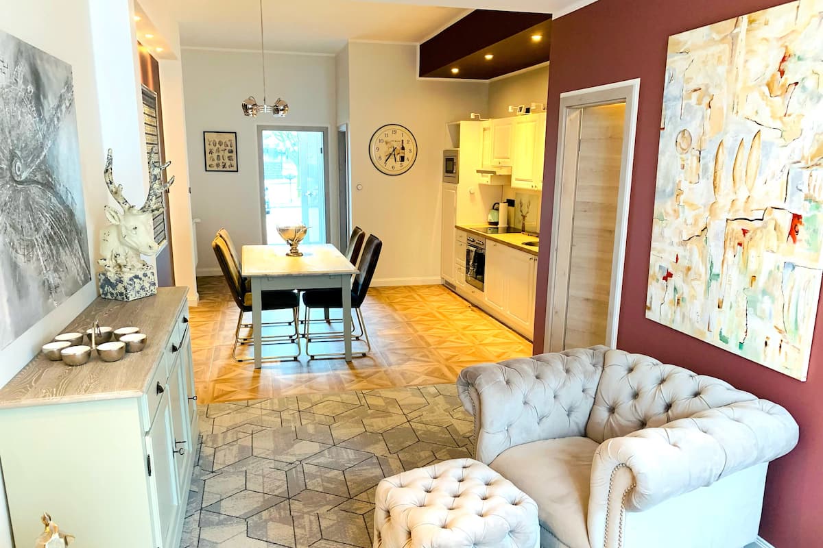 A modern living room with a tufted sofa and ottoman, a dining area with a table and chairs, and a kitchen with white cabinets and a clock on the wall.