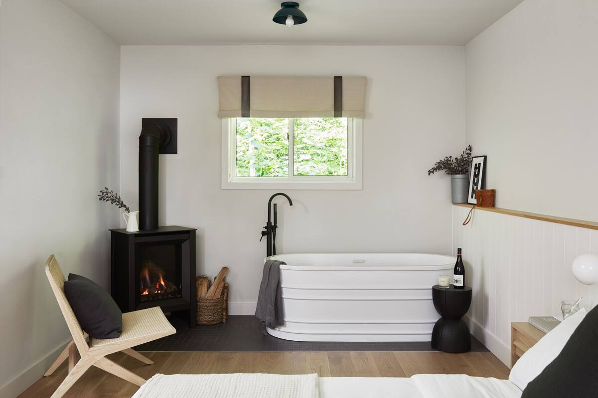 Une salle de bain moderne avec une baignoire blanche, un radiateur noir, une chaise en bois et une fenêtre donnant sur un paysage verdoyant.