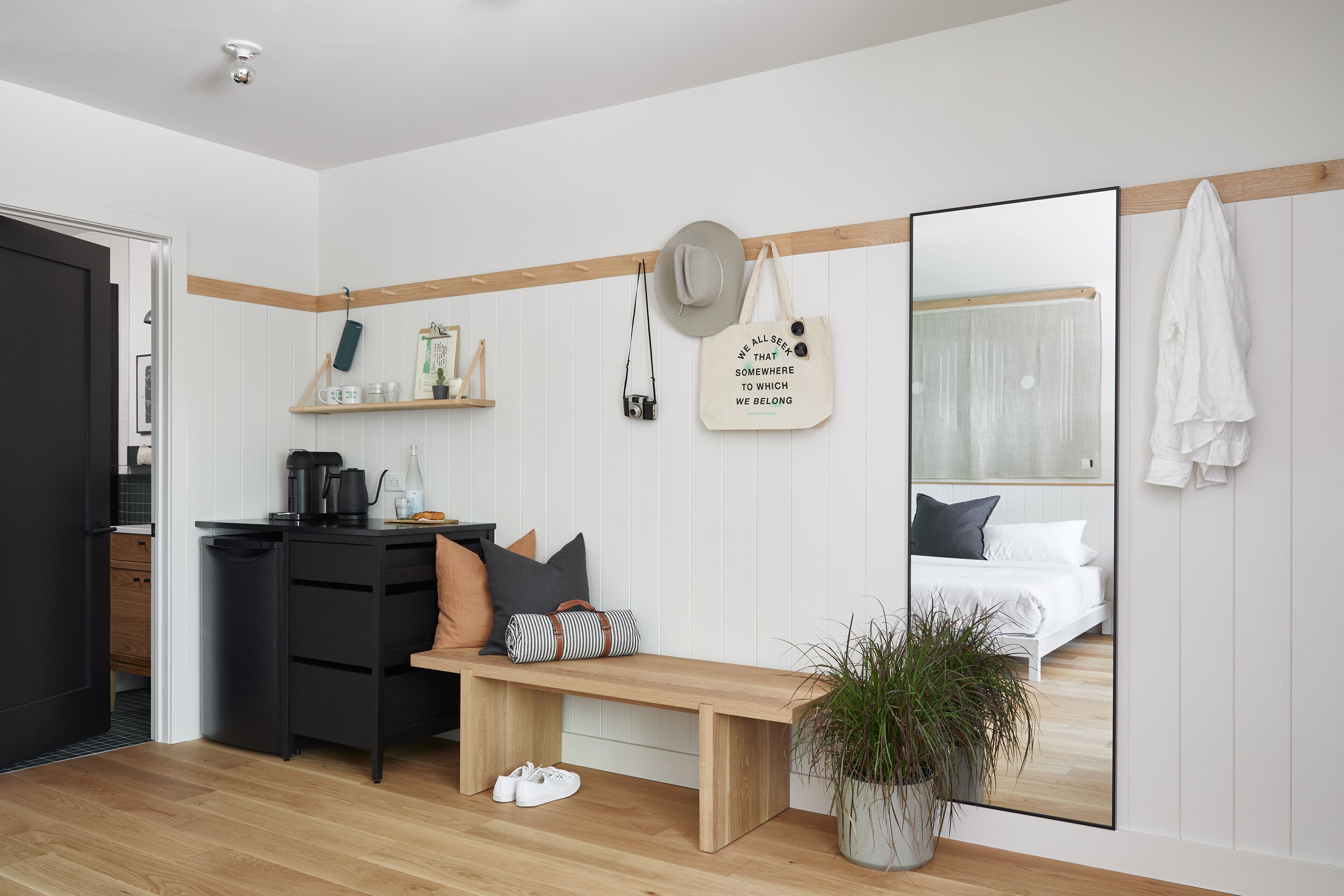 Une chambre à coucher moderne avec un banc en bois, un miroir, une table de chevet avec un appareil photo et une plante en pot.