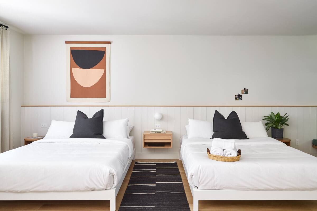 Une chambre d’hôtel avec deux lits, une œuvre d’art accrochée au mur, des tables de chevet avec des lampes, une plante en pot et un plateau avec des serviettes.