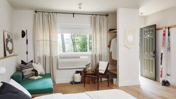 Habitación de hotel con cama, una silla, escritorio, ventana con cortinas, puerta y perchero.