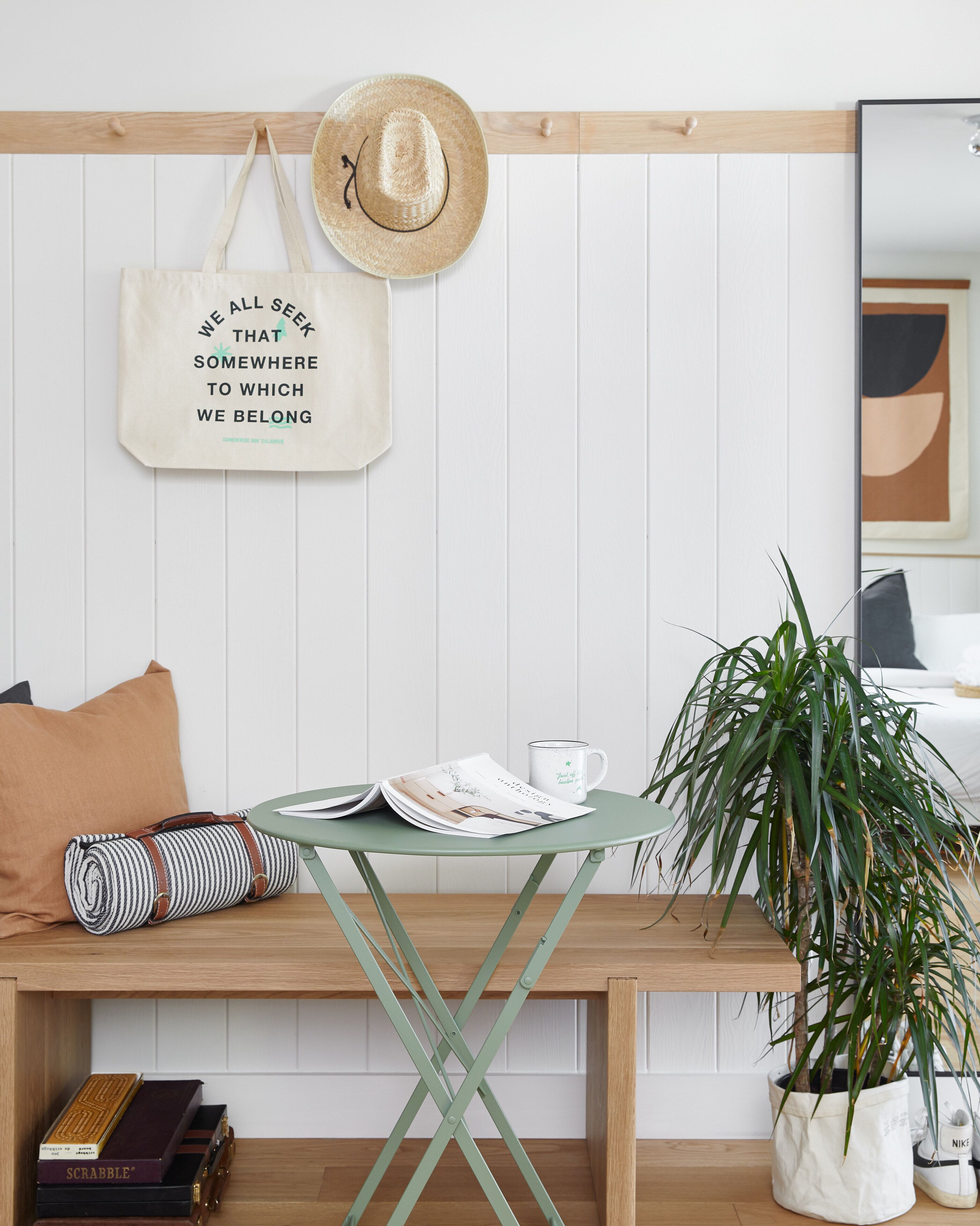 Et pænt møbleret rum med en træbænk, et grønt klapbord, en potteplante, en hat på væggen og et inspirerende citat.