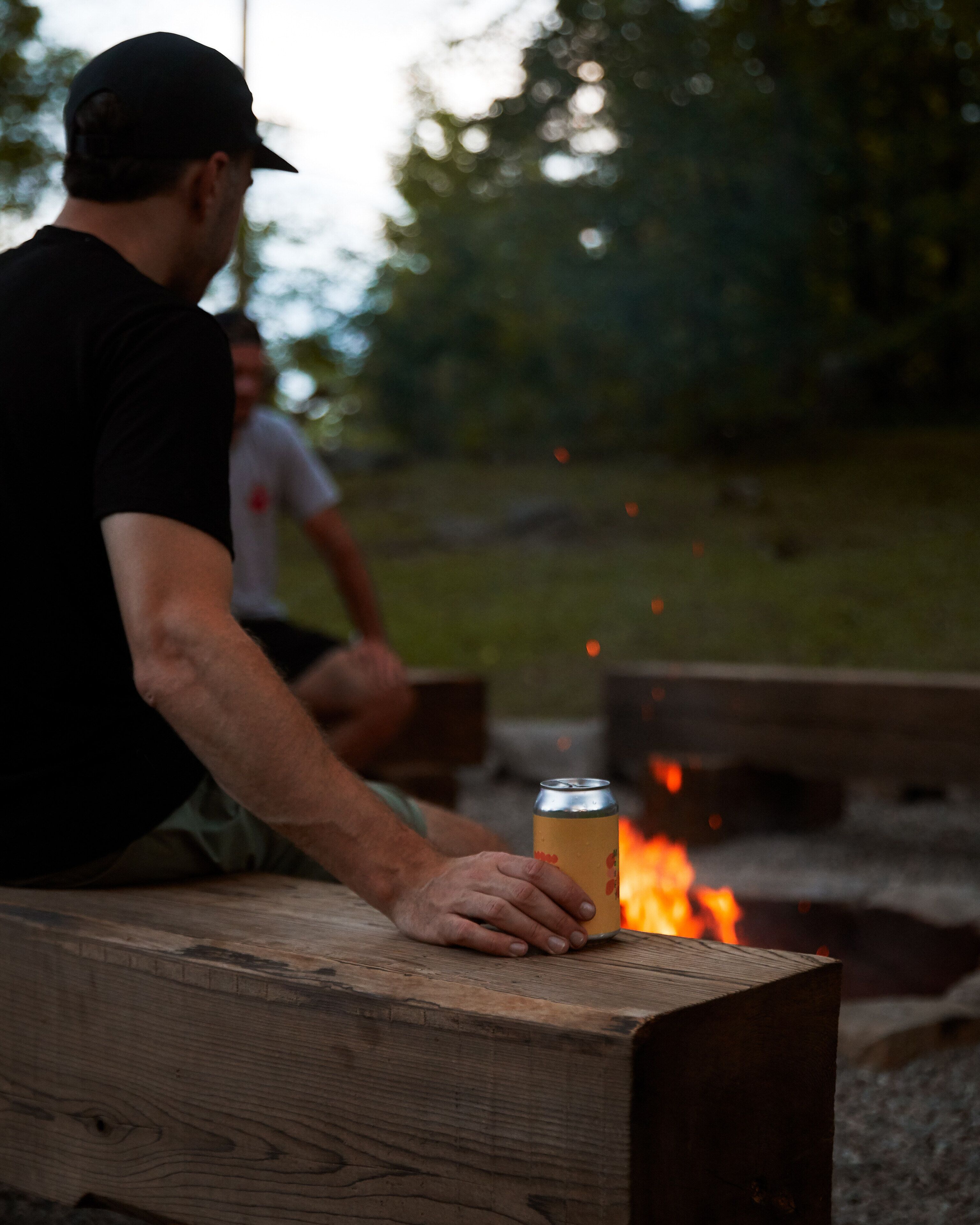 A person sitting at a wooden bench holding a can, with a campfire in the background.