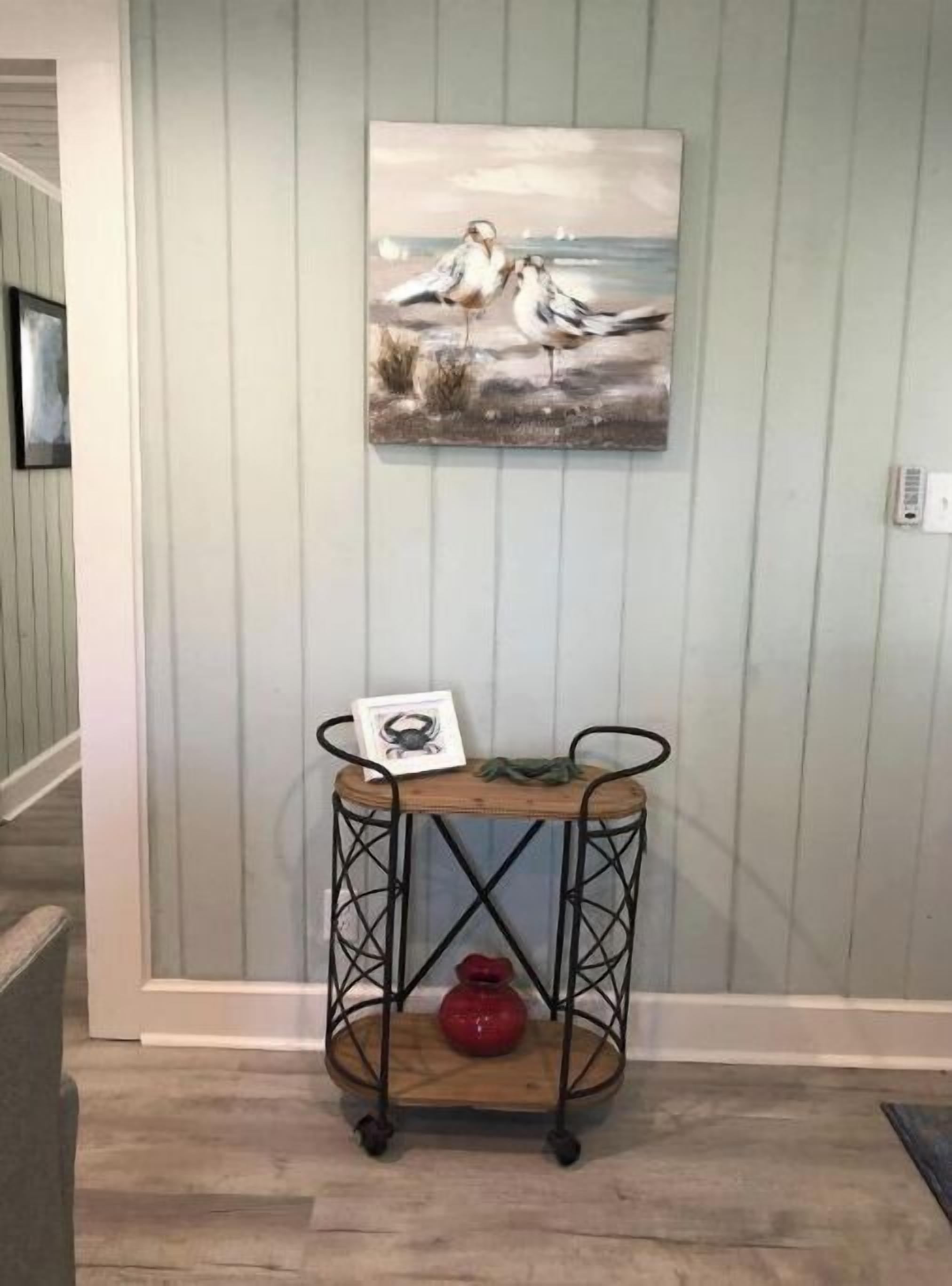 Um corredor com um quadro de pássaros fixado na parede, uma mesa com estrutura de metal e um vaso, e uma moldura com uma fotografia.