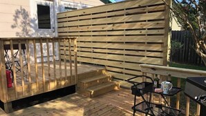 Una terraza de madera con barandilla, una pequeña mesa con sillas y una parrilla para barbacoa.