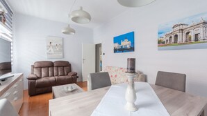 Una sala moderna con un sofá, una mesa de comedor con mantel blanco y una vela decorativa. Hay luces empotradas en la pared y un cuadro de un edificio.