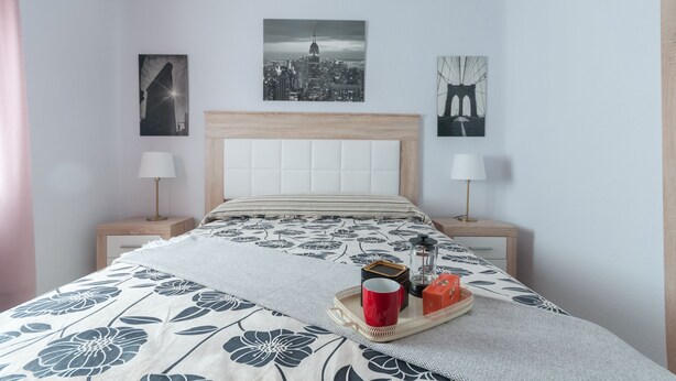 Un dormitorio ordenado con una cama, dos mesitas de noche y una obra de arte con un paisaje urbano en la pared.