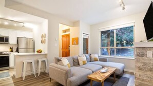 A modern kitchen with white cabinets and stainless steel appliances, a gray sectional sofa with cushions, a wooden coffee table, and a large window with a view of trees.