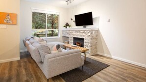 A modern living room with a stone fireplace, a flat-screen TV, a sofa, and a large window offering a view of the outdoors.