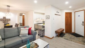 A modern living room with a grey sofa, a coffee table, and a bench. A kitchen is visible in the background with a dining area. - NEW! 2BR Townhouse: Private HotTub, Free Parking (Whistler)