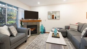 A modern living room with a fireplace, two grey sofas, a coffee table, and a large flat-screen TV mounted above the fireplace. - NEW! 2BR Townhouse: Private HotTub, Free Parking (Whistler)