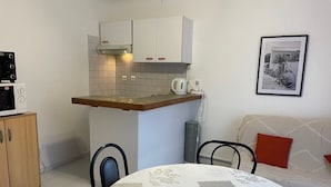 Een kleine keuken met witte kastjes, een magnetron en een waterkoker. Er is een eettafel met stoelen en een bank met een kussen.