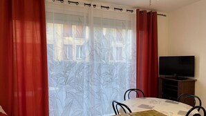 Een eetgedeelte met een tafel en stoelen, een televisie op een houten dressoir en rode gordijnen met een bladmotief.