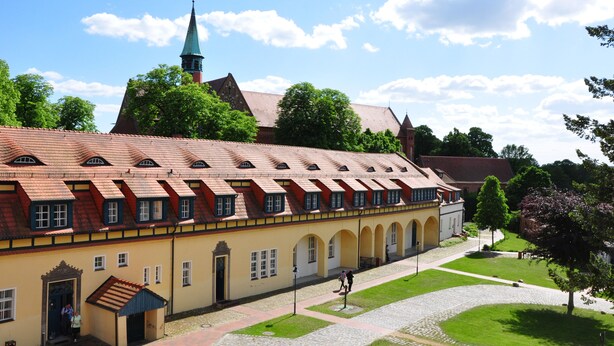 A historic building with a long corridor, arched windows, and a church with a spire in the background. - Zentrum-Kloster-Lehnin (Kloster Lehnin)