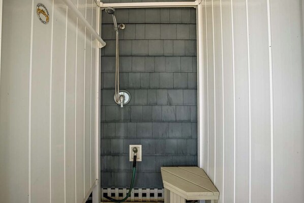 Un petit espace douche fermé, doté d’un pommeau de douche et d’un tuyau en métal, d’un banc blanc et d’un mur blanc avec un lambris vertical.